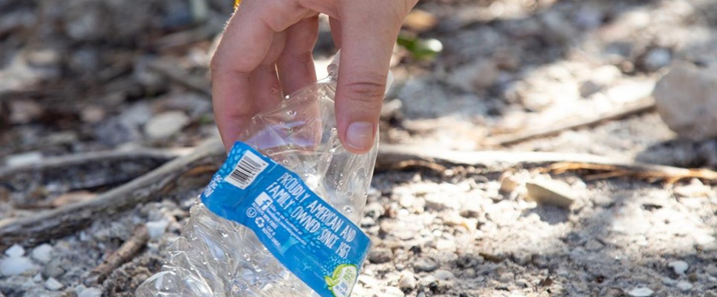 A hand reaches down to pick up a crumpled plastic water bottle.