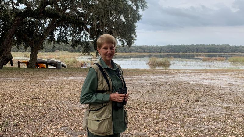 a woman stands beside a lake with binoculars.