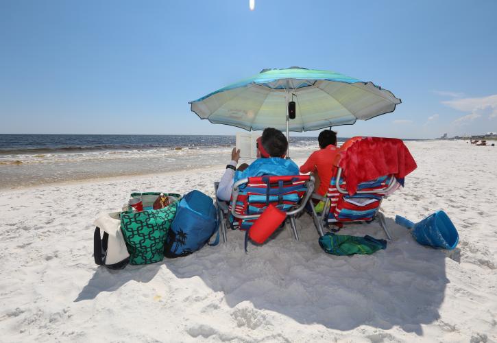 Couple sit under the shade of a beach umbrella reading a book.